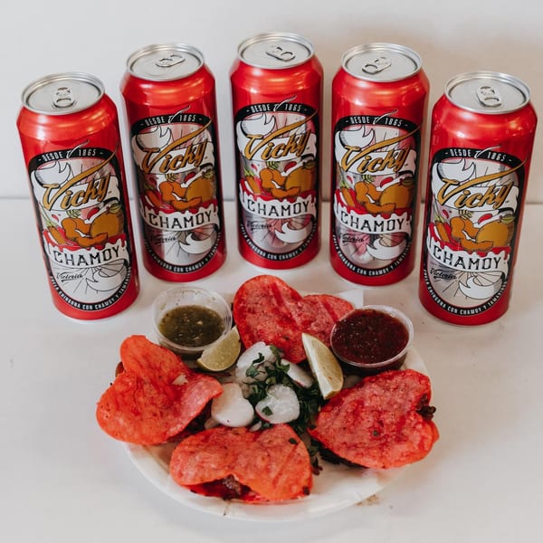 Five Vicky Chamoy beer cans stand behind a plate of heart-shaped red tortillas with limes and sauces.