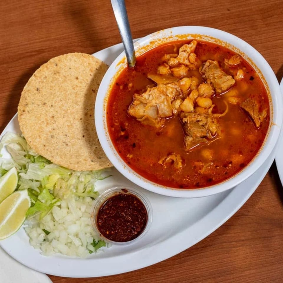 A bowl of Los 4 Vientos Carniceria's pozole with a side of tostada, lettuce, onions, lime, and salsa.