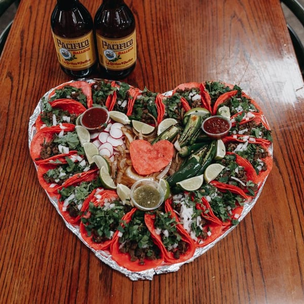 Heart-shaped platter of Los 4 Vientos tacos with red tortillas, limes, radishes, and two Pacifico beers.