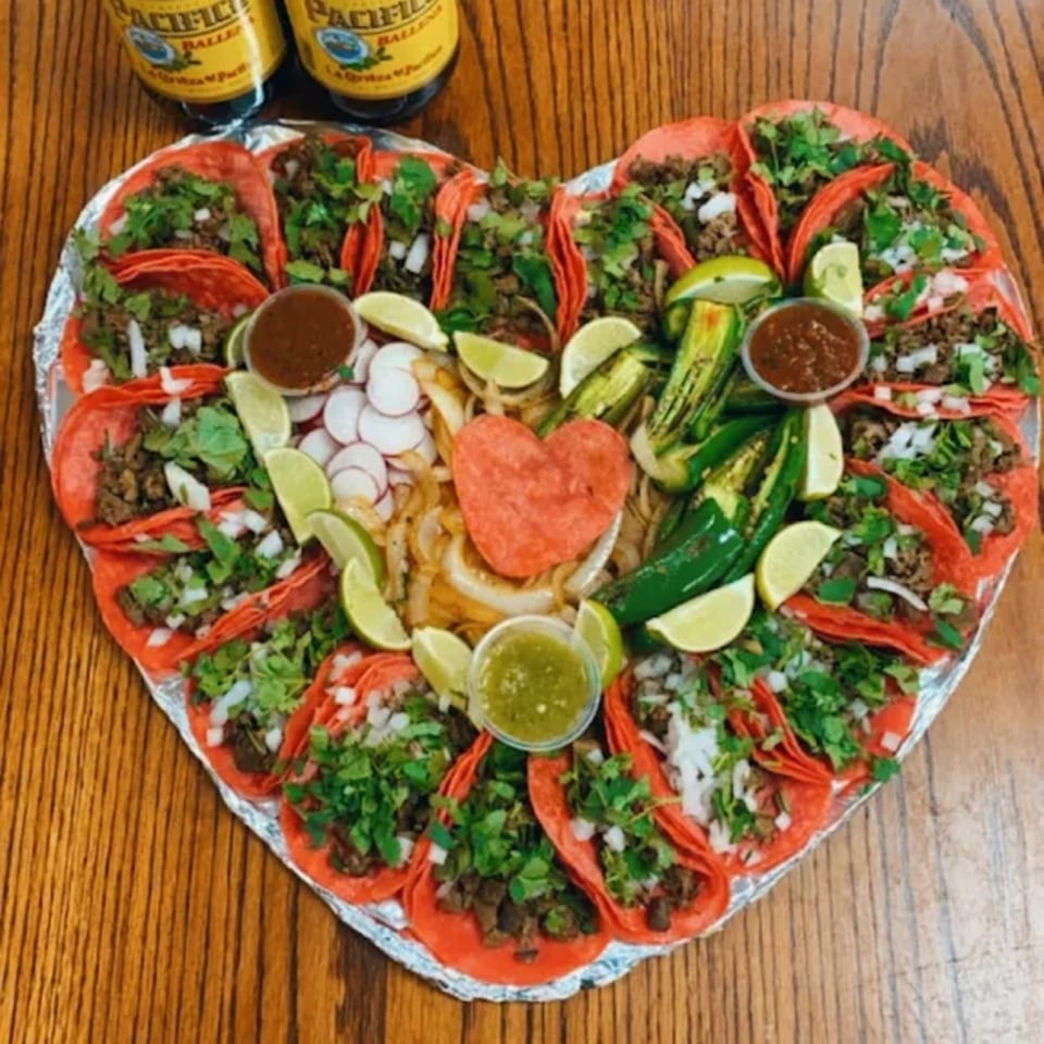 Heart-shaped platter of Los 4 Vientos Carniceria tacos with red tortillas, limes, jalapeños, and sauces.