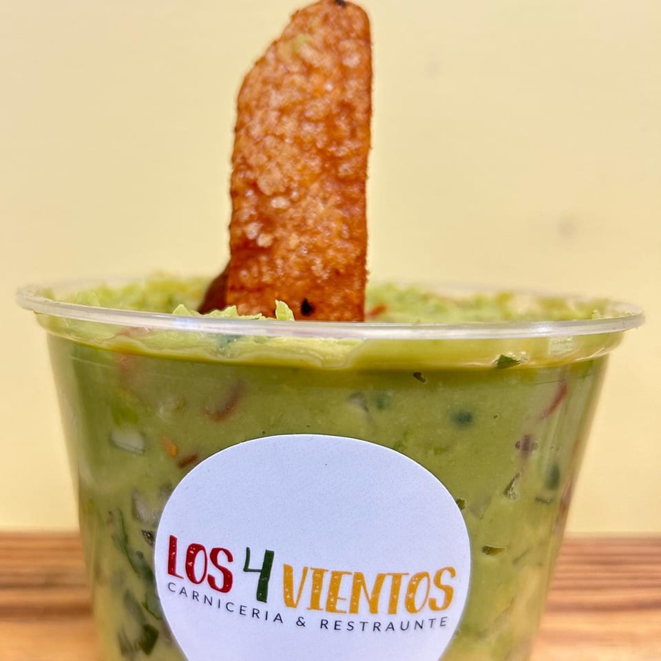 A clear plastic cup of guacamole with a single tortilla chip, branded with Los 4 Vientos logo.