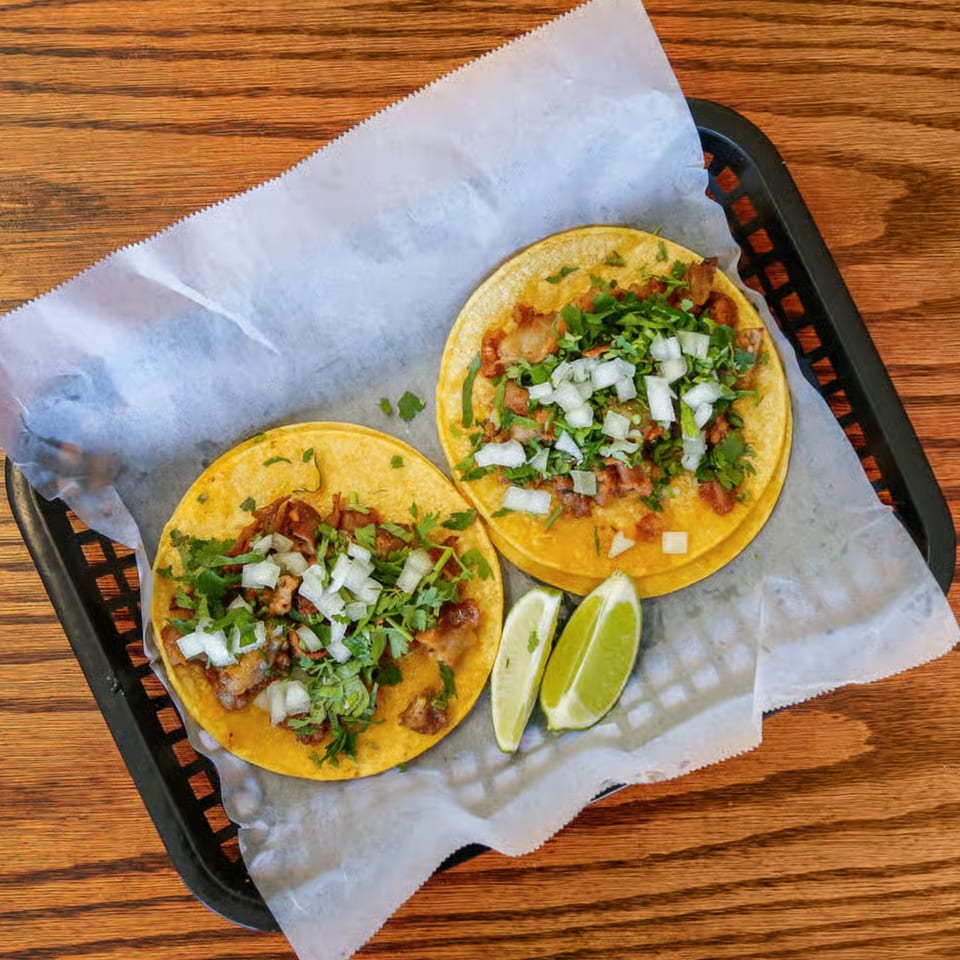 Two Los 4 Vientos Carniceria tacos with meat, cilantro, onions, and lime wedges in a basket.